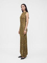 Femme debout portant une longue robe sans manches à imprimé serpent marron et jaune, avec des chaussures pointues noires et un bracelet argenté sur fond uni.