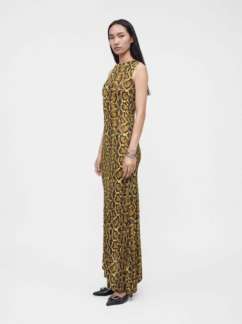 Femme debout portant une longue robe sans manches à imprimé serpent marron et jaune, avec des chaussures pointues noires et un bracelet argenté sur fond uni.
