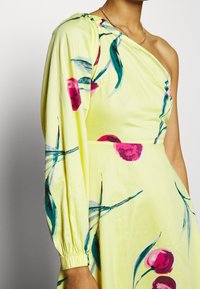 Žena v žlutých šatech na jedno rameno s dlouhým rukávem, zdobených vzorem třešní a listů, a zlatým řetízkovým náhrdelníkem.