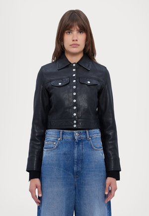 Woman wearing a buttoned black leather jacket with front pockets and loose blue denim jeans, standing against a plain white background.