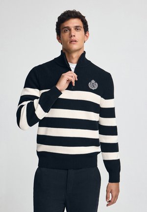 Zwart-wit gestreepte pullover trui met een halve ritssluiting, geribbelde textuur en geborduurd embleem op de borst.