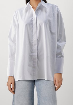 Vrouw die een loszittend wit overhemd met knopen en lichtblauwe spijkerbroek draagt, staande met ontspannen armen langs het lichaam tegen een effen achtergrond.