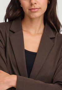 Blazer marrone con una vestibilità sartoriale, revers a punta e un tessuto leggero. Indossato su una maglietta nera, mette in risalto un design semplice e pulito.