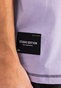 Tricou scurt cu mânecă, de culoare lila, cu o etichetă din material negru. Eticheta conține text alb "STUDIO EDITION" și "ALPHA INDUSTRIES INC."