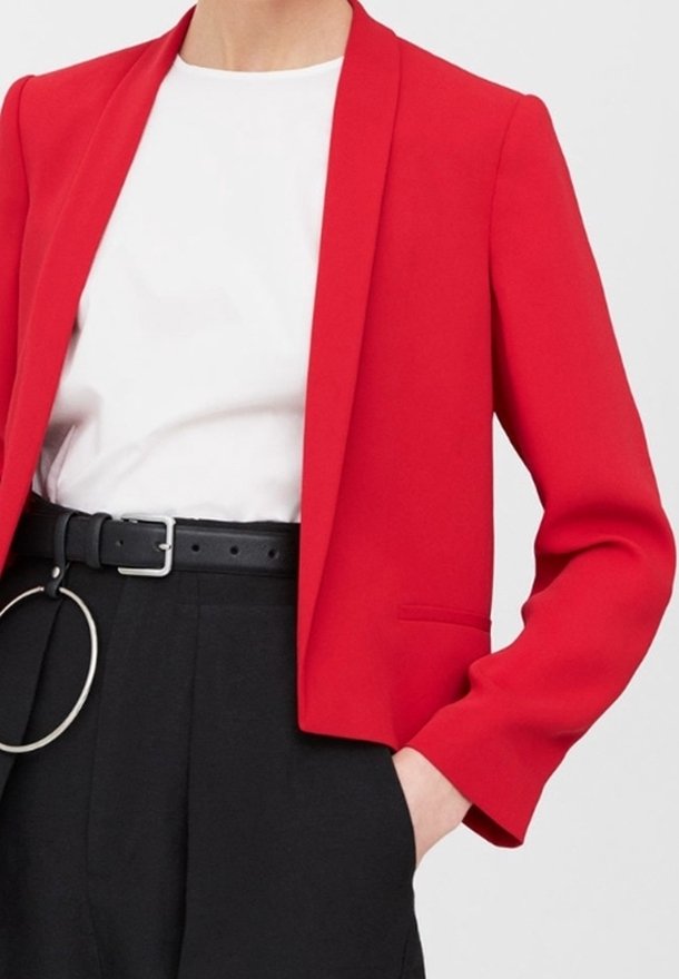 Personne portant un blazer rouge vif sur une chemise blanche, pantalon noir taille haute avec ceinture noire et détail anneau argenté, main dans la poche.