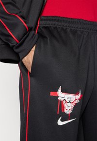 Μαύρο αθλητικό παντελόνι με κόκκινες ρίγες, με εικόνα κεφαλής ταύρου και το λογό τυπο της Nike. Κατασκευασμένο από λείο, ελαστικό ύφασμα.