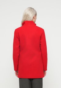 Rdeča plašč iz volnene mešanice z visokim ovratnikom in dolgimi rokavi. Gladka tekstura in prilagojen kroj, ki se konča do sredine stegna. Zadnji pogled.