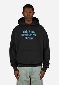 Čierna mikina s kapucňou vyrobená z bavlneného zmesi, na prednej strane s modrým textom "Táto pieseň mi pripomína teba". Obsahuje klokaní vrecko.