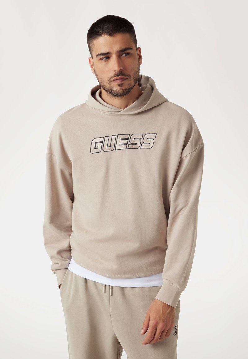 Beige színű pamut kapucnis pulóver, állítható zsinórral, az elején jól látható hímzett "GUESS" logóval. Laza szabás és lágy tapintás jellemzi.