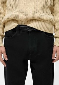 Črne jeans hlače iz denima z stranskimi žepi, v kombinaciji s svetlo bež pletenim puloverjem z ribanim robom in teksturo. Vidna črna pasovina.