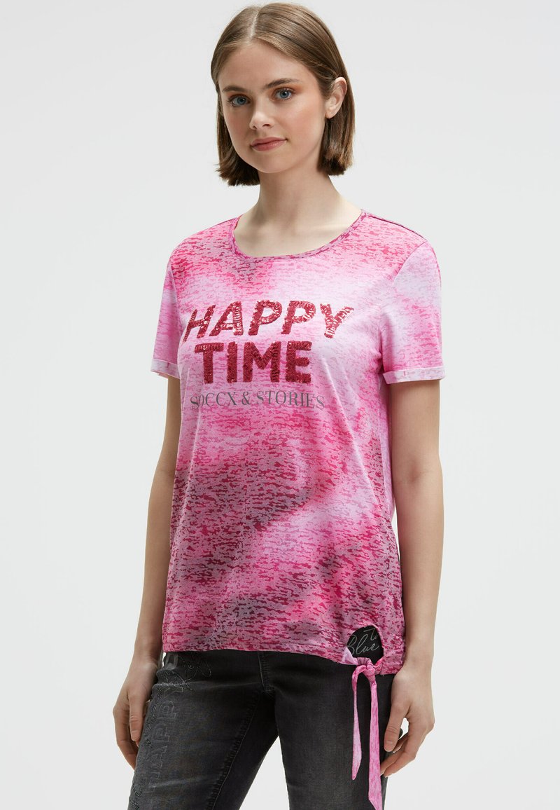 Young woman wearing a pink patterned t-shirt with "HAPPY TIME" text in sequins, paired with dark jeans, standing against plain background.