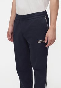Άνδρας που φοράει σκούρο μπλε φόρμα EA7 Emporio Armani με ελαστική μέση και λευκές ρίγες στα πλάγια σε λευκό φόντο.