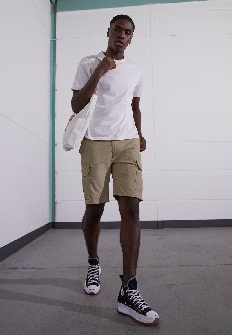 T-shirt blanc à manches courtes, shorts cargo kaki avec poches et baskets montantes noires avec accents blancs et semelles en caoutchouc. Sac fourre-tout en coton.