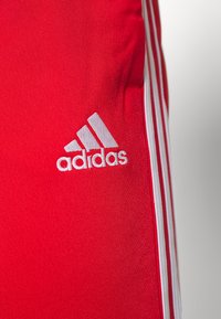 Rdeče atletskih kratkih hlač iz gladkega materiala z belim logotipom Adidas in tremi belimi črtami ob straneh.