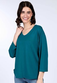 Türkisfarbener Langarmoberteil mit V-Ausschnitt, lockerer Passform und weicher Textur. Mit einem abgerundeten Saum und seitlichen Schlitzen für mehr Komfort. Kombiniert mit Jeans.
