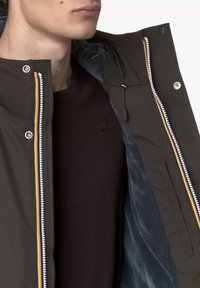 Waterdichte jas in donkerolijfgroen met een contrasterende voering, kleurrijke ritssluitingen en een capuchon met trekkoord. Bruine trui eronder.