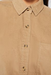Gros plan d'une chemise beige en velours côtelé avec des boutons marron et une poche poitrine avant.