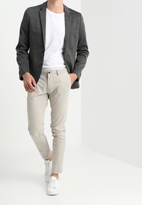 Uomo che cammina indossando un blazer grigio scuro, una t-shirt bianca, pantaloni beige e sneakers bianche su uno sfondo chiaro e uniforme.