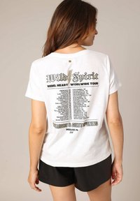 Witte katoenen t-shirt met een zwart en gouden grafisch ontwerp op de rug dat een concerttourlijst weergeeft, korte mouwen en een losse pasvorm.