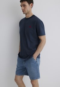 Marineblaues Baumwoll-T-Shirt mit rundem Halsausschnitt und kleinem roten Logo auf der Brust, kombiniert mit hellblauen Denim-Shorts mit Seitentaschen.