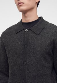 Gros plan d'une personne portant un pull boutonné texturé gris foncé par-dessus un t-shirt noir à col rond, avec un col visible et quatre boutons.