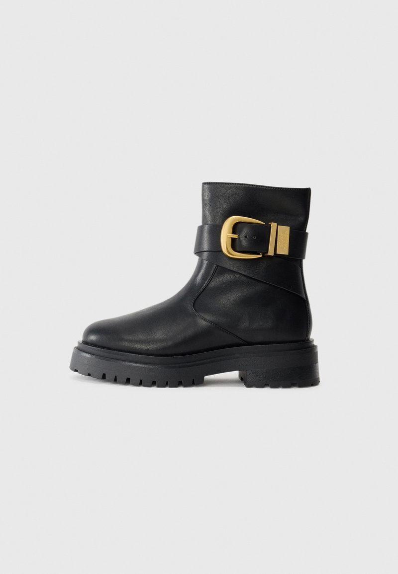Bottine en cuir noir avec semelle épaisse et large sangle à boucle dorée sur fond gris uni.