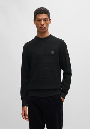 Schwarzer Pullover aus strukturiertem Material, runder Halsausschnitt und Logopatch auf der Brust. Mit engen Bündchen und Saum, kombiniert mit dunklen Cordhosen.
