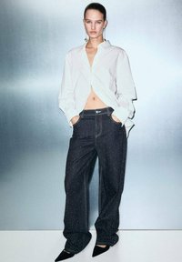 Camisa blanca de manga larga, cortada a la cintura, combinada con jeans oscuros holgados que presentan un sutil patrón moteado, y tacones negros de punta.