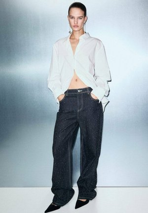 Camisa blanca de manga larga, cortada a la cintura, combinada con jeans oscuros holgados que presentan un sutil patrón moteado, y tacones negros de punta.