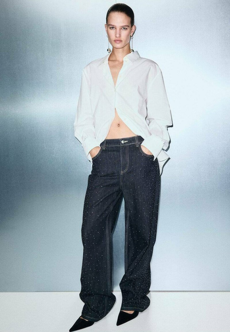 Camisa blanca de manga larga, cortada a la cintura, combinada con jeans oscuros holgados que presentan un sutil patrón moteado, y tacones negros de punta.
