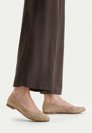 Füße in beigen Wildleder-Ballerinas mit niedrigen Holzabsätzen, kombiniert mit lockeren, dunkelbraunen Hosen bis zum Knöchel.