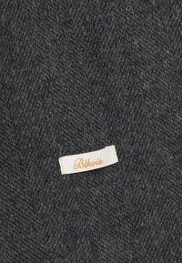 Tissu gris foncé avec une texture en laine, arborant une petite étiquette blanche brodée avec "Blkvis" en script orange.