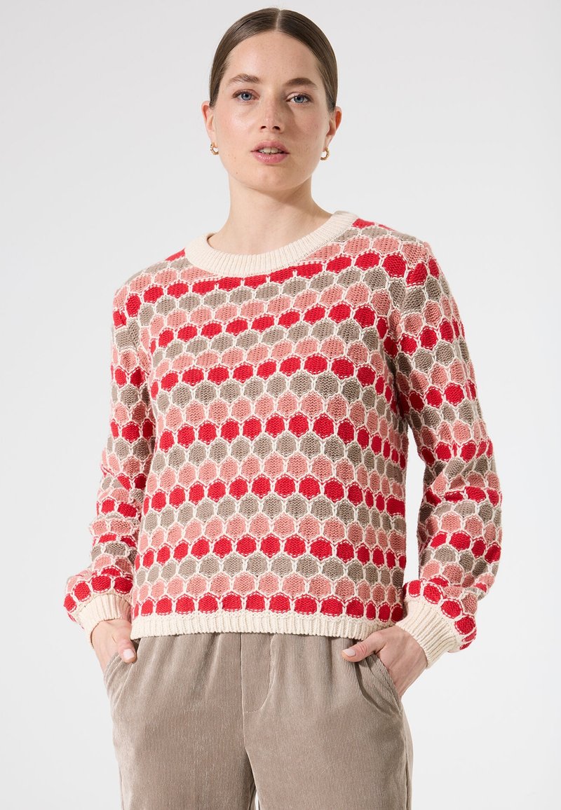 Pull en tricot avec un motif géométrique en rouge, rose, beige et crème. Il a un col rond et des poignets et un ourlet côtelés.