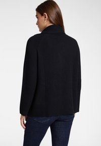 Vrouw van achteren gezien, gekleed in een zwart geribd gebreide trui en donkerblauwe spijkerbroek, voor een effen lichte achtergrond.