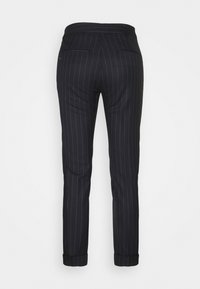 Pantalones negros a medida con rayas blancas verticales finas, con dos bolsillos traseros de ojal y bajos con puños, vistos desde atrás.