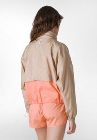Letvægtig, tofarvet jakke med en sandfarvet overdel og lys koral nederdel, der har en høj krave og elastiske manchetter for et tætsiddende look.