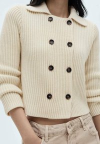 Maglione a coste color crema con colletto, chiusura a doppiopetto con bottoni e maniche a tre quarti. Texture liscia.