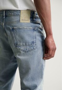 Achteraanzicht van een persoon die lichte blauwe denim jeans draagt met het merklabel "DSTREZZED" boven de achterzak en de zichtbare rechterarm.
