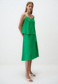 Haut en soie vert, sans manches, superposé à une jupe verte arrivant au genou avec une fente sur le côté. La jupe a un design fluide et une texture douce.