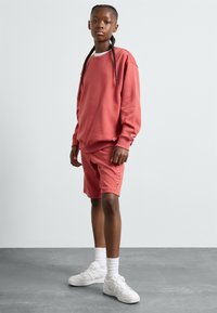 Korallfarget sweatshirt med senkede skuldre, kombinert med matchende shorts som har sideknapper. Brukt med hvite sneakers og ankelstrømper.