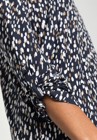 Marinebluse mit einem weißen und beigen Punktmuster. Überrollte Ärmel mit einem Knopfdetail; weicher Stoff und lockere Passform.
