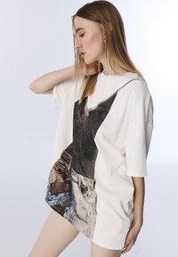 Oversized white T-shirt featuring a graphic print of denim shorts with pockets. Soft cotton fabric, short sleeves, relaxed fit.