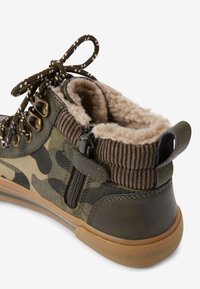 Olijfgroene enkellaarsjes met camouflagepatroon, voorzien van een zachte, beige fleecevoering, bruine ribbelafwerking en een zijrits voor makkelijk aan- en uittrekken.