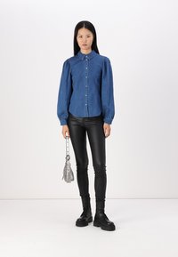Camisa de mezclilla azul con cuello, mangas abullonadas y cierre de botones, combinada con leggings de cuero negro y botas negras de suela gruesa. Accesorio de bolso plateado.