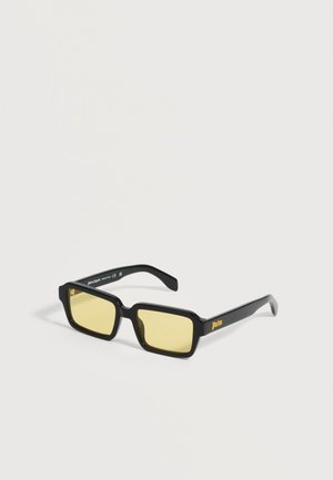 Lunettes de soleil rectangulaires noires avec des verres teintés jaunes et le logo "Palm" sur la branche, posées sur une surface unie gris clair.