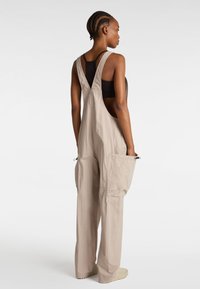 Beige Latzhosen mit weitem Bein, verstellbaren Trägern und seitlichen Taschen, aus leichtem Stoff gefertigt, mit strukturierter Oberfläche und lockerer Passform.
