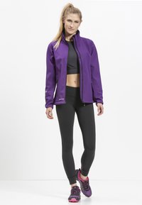 Lilla zip-up jakke med høj krave, parret med en sort sports-bh og sorte leggings. Fodtøjet har lyserøde detaljer og en tekstureret sål.