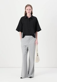 Camisa preta de botão com mangas curtas, combinada com calças largas de cintura alta, cinza claro. O modelo segura uma pequena bolsa branca.