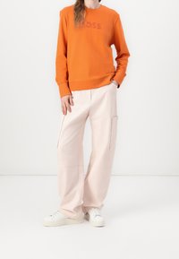 Orange sweatshirt with "BOSS" logo, paired with light pink wide-leg pants featuring side pockets, and white sneakers on a plain background.