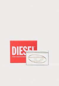 Sidabrinė kortelių laikiklis su ryškiu ovalo formos logotipu, lygiu paviršiumi ir siūlėmis kraštuose, padėtas šalia raudonos Diesel prekės ženklo dėžutės.